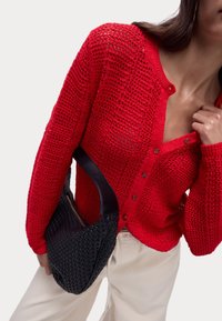 Cardigan rosso a maglia con scollo a V, dettagli sui bottoni e un motivo a trama aperta, abbinato a una borsa a spalla nera intrecciata con manico curvo.