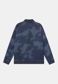 Marinblå långärmad jacka med ett kamouflage-mönster, ribbad krage och ärmslut, samt röd accentstickning längs axlarna. Slät tygtextur.