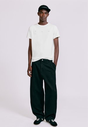 Ung mand iført en hvid T-shirt, sorte vidde bukser, sorte sneakers og en sort kasket, stående mod en ensfarvet lys baggrund.