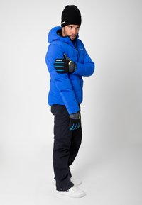 Blue insulated jacket with a hood, black gloves featuring blue accents, dark pants, and white shoes. Model facing slightly to the side.