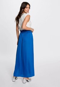 Gonna maxi di un brillante blu, realizzata in un materiale leggero con una texture plissettata. Abbinata a una maglietta bianca senza maniche e sandali con tacco quadrato argentati.