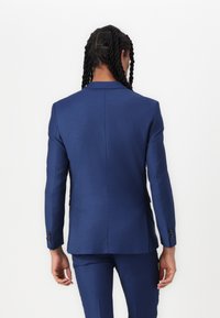 Veste de costume bleu marine sur mesure, design structuré avec une fente arrière, revers cranté et poignets boutonnés, fabriquée en tissu texturé.