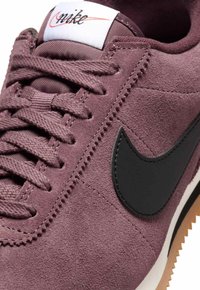 Close-up van een paarse suède Nike sneaker met een zwarte swoosh, veters en een wit label op de tong met het oranje en zwarte Nike logo.