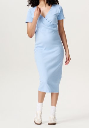 Jasnoniebieska sukienka do kolan z kopertowym dekoltem w serek i krótkimi rękawami, wykonana z gładkiej tkaniny. Noszona z białymi skarpetkami i kremowymi trampkami.