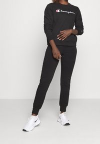 Ensemble sweat-shirt noir et joggers, avec un logo sur la poitrine, des poignets et ourlets aux chevilles côtelés, associé à des chaussures de sport blanches.