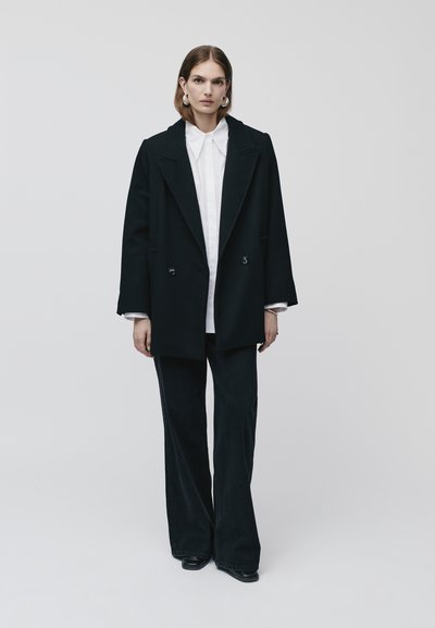 Cappotto doppiopetto nero con revers, indossato su una camicia bianca, abbinato a pantaloni in denim scuri a gamba larga e scarpe nere.