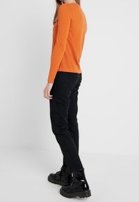 Frau in einem orangefarbenen Langarmshirt, schwarzen geraden Jeans und glänzenden schwarzen Schnürstiefeln, die seitlich auf einem einfarbigen Hintergrund steht.
