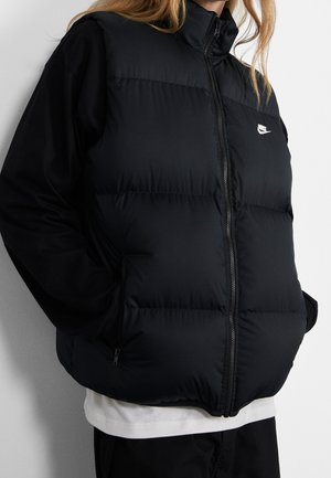 Personne portant un gilet matelassé noir Nike sur un t-shirt à manches longues noir et un sous-vêtement blanc, debout les mains dans les poches du gilet.