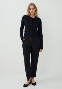 Frau mit blonden Haaren steht vor einem weißen Hintergrund, trägt eine schwarze geknöpfte Strickjacke mit Blumendetails, schwarze Hosen und schwarze Ballerinas.