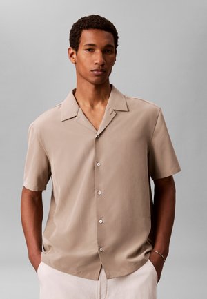 Jeune homme portant une chemise beige à manches courtes boutonnée avec col, debout les mains dans les poches d’un pantalon blanc sur un fond gris uni.