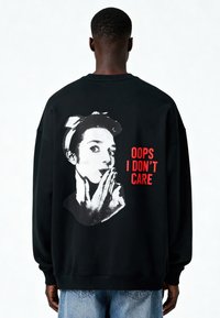 Felpa nera con grafica di una donna con fascia per capelli, stampata in bianco e con testo rosso "OOPS I DON'T CARE" sul retro. Vestibilità comoda.