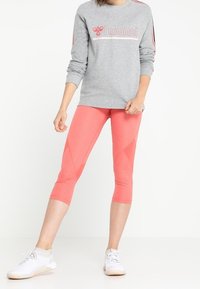 Sudadera gris de manga larga con logo, combinada con leggins capri de color rosa coral con patrones geométricos y zapatillas deportivas blancas.