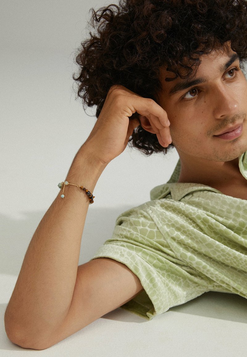 Chemise à motifs verts, texture superposée. Bras orné d'un bracelet en perles multicolores. Fond neutre, modèle avec des cheveux bouclés.