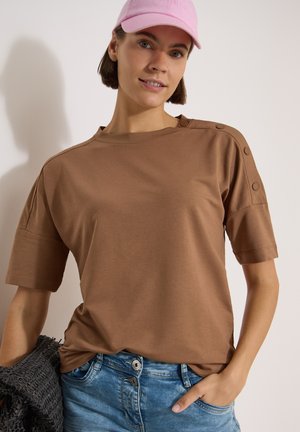 Jeune femme portant un haut marron boutonné aux épaules, jean bleu, casquette rose, tenant un sac noir texturé, souriant légèrement.