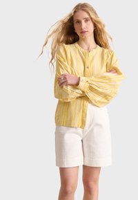Camicia gialla a righe con maniche a palloncino, realizzata in tessuto leggero. Abbinata a pantaloni corti di lino bianchi, su uno sfondo neutro.