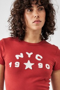 Červené priliehavé tričko s krátkymi rukávmi, ktoré má biele písmo "NYC 1990" a dizajn hviezdy na prednej strane. Hladká bavlnená textúra.