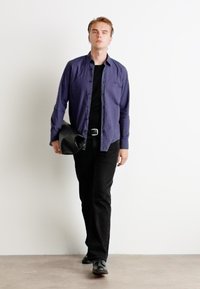 O persoană îmbrăcată cu o cămașă albastră cu nasturi peste un tricou negru, pantaloni negri și pantofi negri, ținând o geantă din piele neagră în timp ce merge.