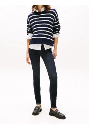 Femme portant un pull rayé bleu marine et blanc sur une chemise blanche, un jean skinny foncé et des mocassins en cuir noir, debout sur un fond blanc.