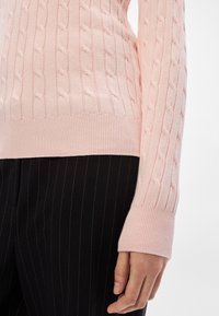 Gaiši rozā džempers ar kabeļa veida adījumu un ribotām aprocēm, ar teksturētu rakstu. Kombinēts ar melnām biksēm ar smalkām svītrām kontrastam.