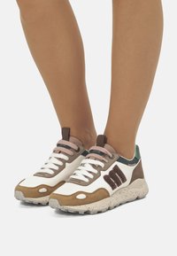 Sneakers con una tomaia in pelle bianca e marrone, caratterizzati da lacci rosa, accenti verdi e una suola beige maculata. Dettagli testurizzati notevoli sul lato.