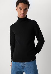 Μαύρο πουλόβερ με ψηλό λαιμό φτιαγμένο από μαλακό ύφασμα, με εφαρμοστό σχέδιο και ραμμένες μανσέτες. Συνδυασμένο με ανοιχτό μπλε τζιν.