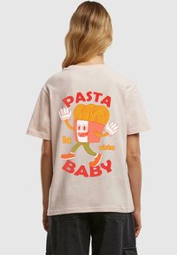 Růžové tričko s krátkým rukávem a volným střihem, na kterém je kreslená postavička se spaghetti vlasy, červený nápis "PASTA BABY" a zelené kalhoty.