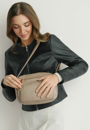 Kobieta w czarnej skórzanej kurtce rozsuwa zamek beżowej torby na ramię na białym tle.