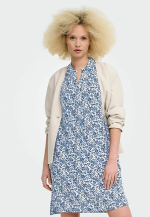 Kvinde med krøllet blondt hår iført guld øreringe i hoop-stil, en beige blazer med op-rullede ærmer og en blå kjole med blomsterprint.