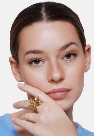 Junge Frau mit braunen Haaren und grünen Augen, die ihr Kinn auf eine Hand mit einem großen goldfarbenen blumenförmigen Ring stützt, trägt ein hellblaues Oberteil.