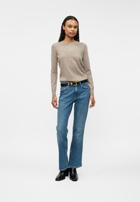 Pull en tricot brun clair, jean droit bleu, ceinture noire avec boucle en or, bottines noires, modèle usé se tenant contre un fond blanc.