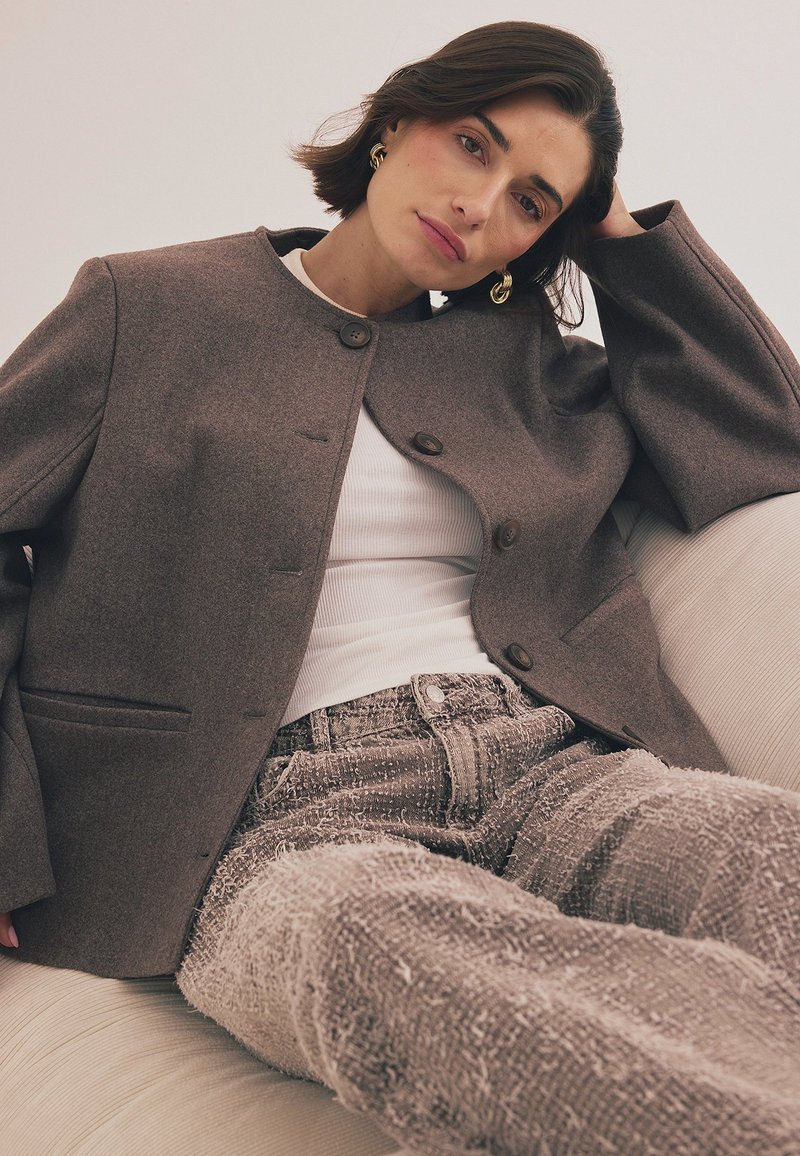Rjav volnen blazer z okroglim izrezom in gumbno zaponko, v kombinaciji z teksturiranimi sivimi jeans hlačami. Gladki materiali in značilne detajle šivanja.