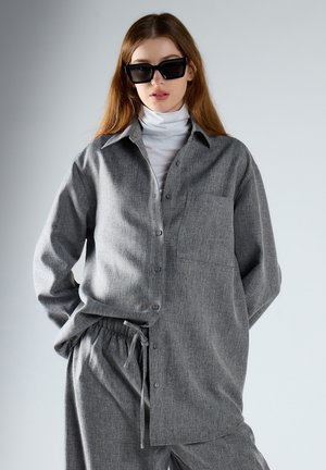 Jonge vrouw die een oversized grijs overhemd met knopen draagt, bijpassende broek met trekkoord, witte coltrui en grote zwarte zonnebril.