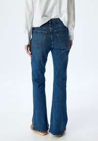 Person, der eine blaue Denim-Jeans im Schlagstil und ein weißes langärmliges Hemd trägt, steht mit entspannten Händen an den Seiten vor einem weißen Hintergrund.