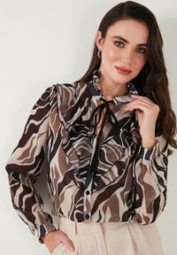 Blusa trasparente con colletto e parte anteriore a volant, caratterizzata da un motivo astratto marrone e beige. Dettaglio con cravatta nera e chiusure a bottone lungo la parte anteriore.