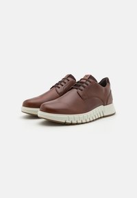 Brown leather sneakers with round toes, lace-up design, and white flexible rubber soles featuring textured detailing along the side.