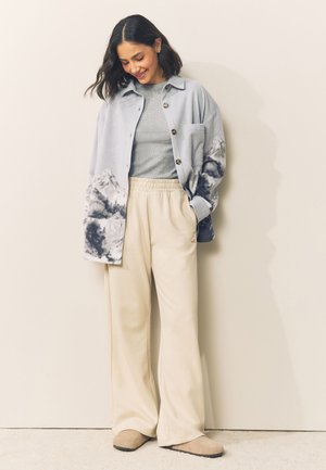 Femme aux cheveux mi-longs foncés portant un t-shirt tie-dye bleu clair, un haut gris, un pantalon beige à larges jambes et des chaussures beiges sans lacets, souriante et regardant vers le bas.