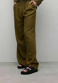 Olijfgroene, wijde broek gemaakt van een gestructureerde stof, gecombineerd met zwarte sandalen met gespen en een dikke zool, tentoongesteld op een neutrale achtergrond.