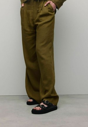 Olijfgroene, wijde broek gemaakt van een gestructureerde stof, gecombineerd met zwarte sandalen met gespen en een dikke zool, tentoongesteld op een neutrale achtergrond.