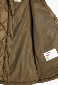 Olivgrüne gesteppte Jacke mit einem glatten, glänzenden Innenfutter, ausgestattet mit Druckknöpfen und einem Etikett mit Pflegehinweisen auf der rechten Seite.