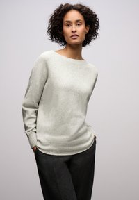 Woman with curly hair wearing a light gray sweater and dark pants, standing with hands in pockets against a plain background.
