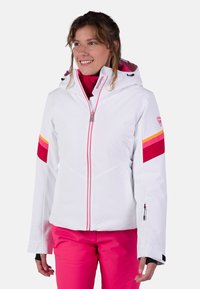 Witte skijas met roze en oranje strepen. Voorzien van een rits aan de voorkant, een capuchon en ritsmanchetten. Gemaakt van een glad, waterdicht materiaal.
