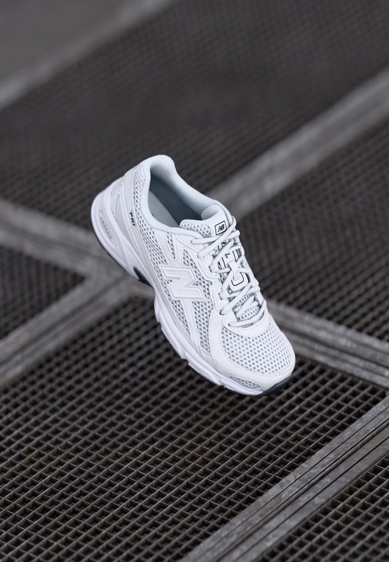 Chaussure de course blanche New Balance avec un design en mesh et des lacets, posée sur une surface en grille métallique sombre.
