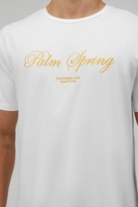 Camiseta blanca de algodón con texto bordado en amarillo "Palm Spring" y "California Love Eighty Five" debajo, de manga corta y con cuello redondo.