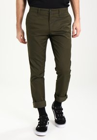 Olivgröna, slim fit chinos med knappstängning, uppvikta benslut och fickor. Kombinerat med svarta sneakers med vita detaljer.