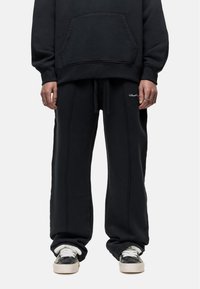 Zwarte katoen sweatshirt met een voorzak en bijpassende losse sweatpants. Beide hebben witte stiksels en minimale branding. Zwarte sneakers met witte accenten.