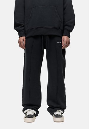 Black cotton sweatshirt with a front pocket and matching loose-fit sweatpants. Both feature white stitching and minimal branding. Black sneakers with white accents.