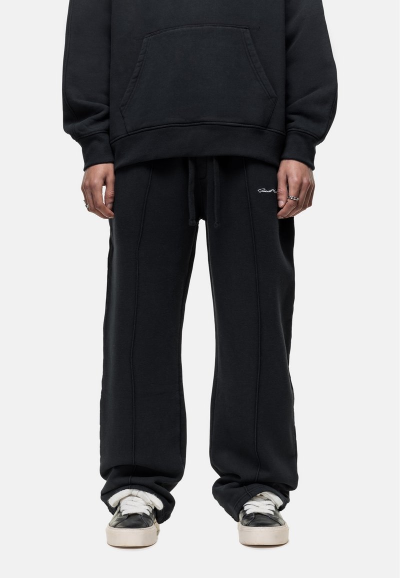 Zwarte katoen sweatshirt met een voorzak en bijpassende losse sweatpants. Beide hebben witte stiksels en minimale branding. Zwarte sneakers met witte accenten.