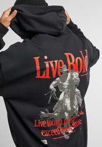 Felpa nera con cappuccio caratterizzata da una grande grafica sulla schiena raffigurante un cowboy a cavallo, con testo rosso e bianco che dice "Vivi audace" e "Vivi forte, resisti".