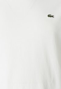 Camiseta blanca de algodón de manga corta y cuello en V, con un pequeño logotipo de cocodrilo bordado en verde en el pecho. Textura suave.