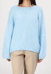 Personne portant un pull ample bleu clair tricoté et un pantalon beige, debout les mains détendues le long du corps devant un fond uni.